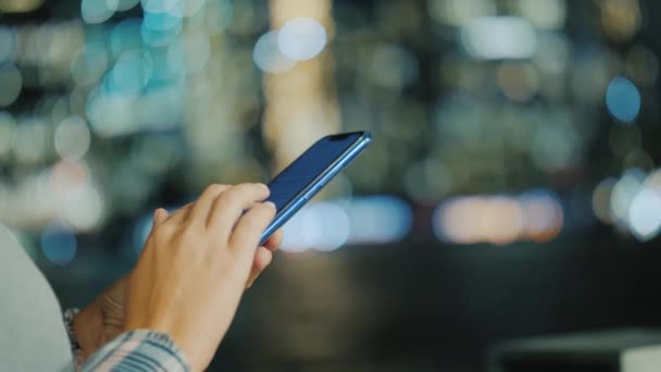 Manos de una mujer joven con un teléfono inteligente en el fondo de las luces divididas de la ciudad — Vídeos de Stock