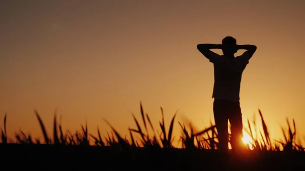 Silueta Un joven disfruta del aire fresco y admira la puesta de sol. Él sostiene sus manos detrás de la parte posterior de su cabeza, mira hacia adelante delante de él —  Fotos de Stock