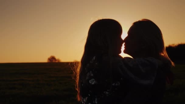 Una mujer besa a su hija tiernamente. El sol al atardecer ilumina maravillosamente sus siluetas — Vídeos de Stock