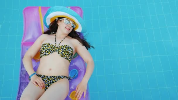 Девушка-туристка в купальнике, отдыхающая в бассейне на надувном матрасе в разноцветной шляпе и солнечных очках . — стоковое видео
