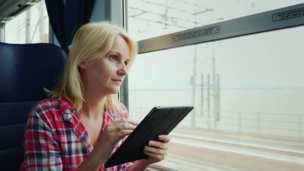 Una joven viaja en tren. Se sienta junto a la ventana, utiliza una tableta digital — Vídeo de stock
