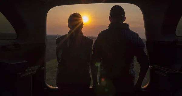 Um jovem casal se senta ao lado do tronco de um carro, admirando o belo pôr do sol em um lugar pitoresco nas montanhas — Fotografia de Stock