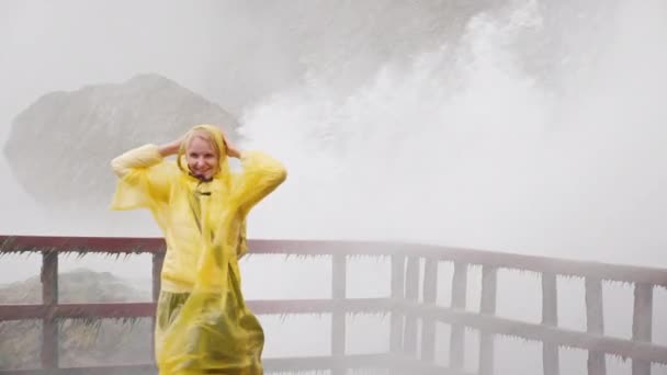 Una mujer con un impermeable amarillo disfruta salpicando agua bajo una gran cascada. Vídeo en cámara lenta — Vídeos de Stock