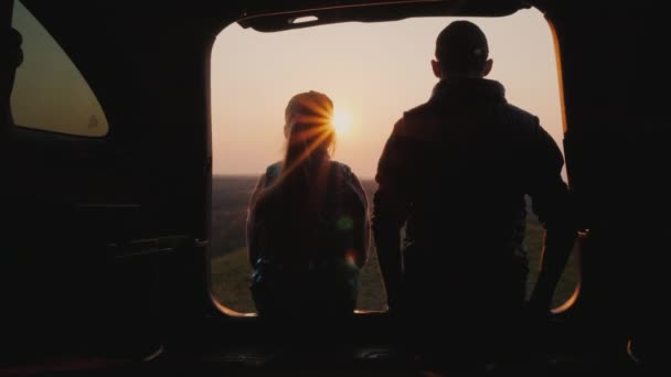 Tonåring med yngre syster sitter i bagageluckan på en bil och tittar på solnedgången — Stockvideo