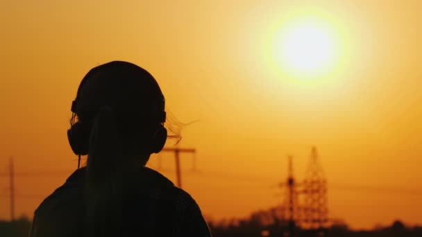 Chica en auriculares escuchando la radio en el fondo del paisaje urbano y hermoso cielo naranja al atardecer — Vídeo de stock