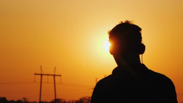 Adolescente in cuffia ascoltando musica, guardando un bel tramonto sulla città, vista da dietro — Video Stock