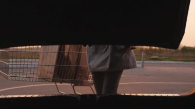 Kadın silueti, bir arabanın bagajını açar, oraya kağıt alışveriş torbaları koyar.