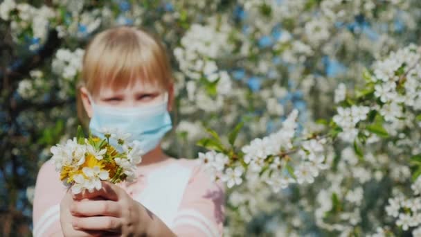 Klein meisje met een boeket bloemen en een masker op de achtergrond van bloeiende bomen — Stockvideo