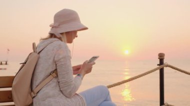 Pembe giyimli şık bir kız akıllı telefondan müzik dinleyip, deniz iskelesinde pembe bir gün batımının ışınlarıyla bankta oturuyor.