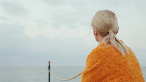 Dreamy girl s'assoit sur la jetée par une matinée fraîche, fouille dans une couverture et écoute de la musique sur un casque — Video