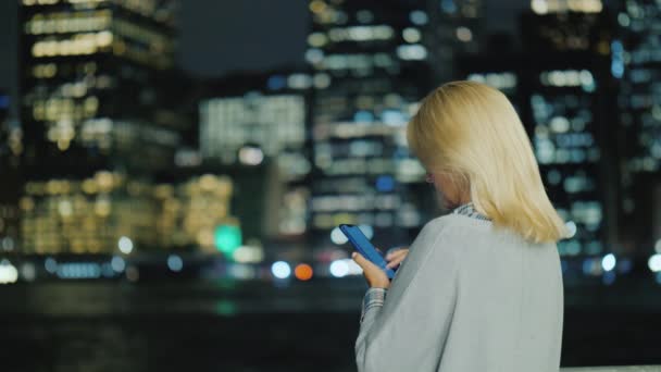Vista trasera de la mujer joven con un teléfono inteligente en el fondo de las luces de una gran metrópolis — Vídeos de Stock