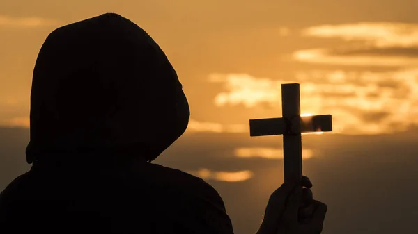 Un moine dans une hotte avec un crucifix dans les mains se tient sur la toile de fond d'un ciel dramatique au coucher du soleil — Photo