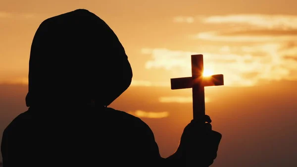 A monk in a hood with a crucifix in his hands stands against the backdrop of a dramatic sky at sunset — Stock Photo, Image