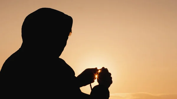 Silhouette d'un homme dans le capot, passant au crible le chapelet au coucher du soleil — Photo