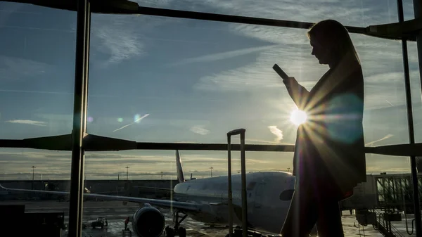 Een vrouwelijke passagier met een telefoon in het terminalvenster van de luchthaven — Stockfoto