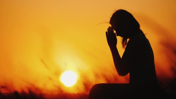 Silhouet van een vrouw die bidt tegen de achtergrond van een oranje hemel en een grote ondergaande zon — Stockvideo