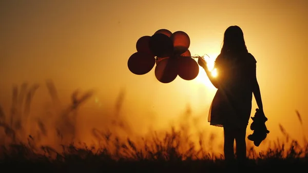 Silhueta de uma menina com balões e um ursinho de pelúcia. Vale o pôr-do-sol. Adeus ao conceito de infância — Fotografia de Stock