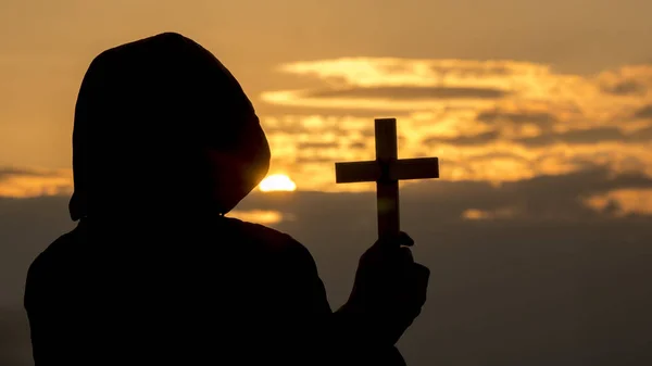 Silhouette of a man in a hood with a crucifix in his hand — Stock Photo, Image