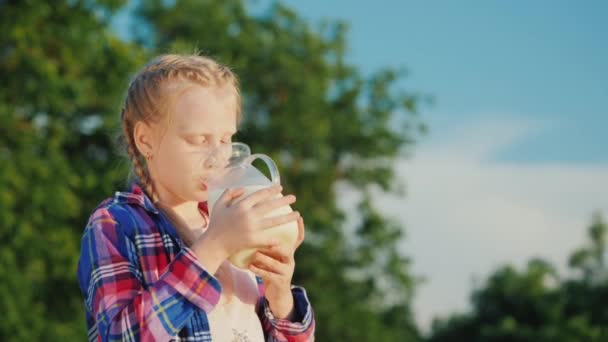 Ein Kind trinkt Milch aus einem Krug vor einem Hintergrund grüner Bäume. frische und gesunde Lebensmittel — Stockvideo