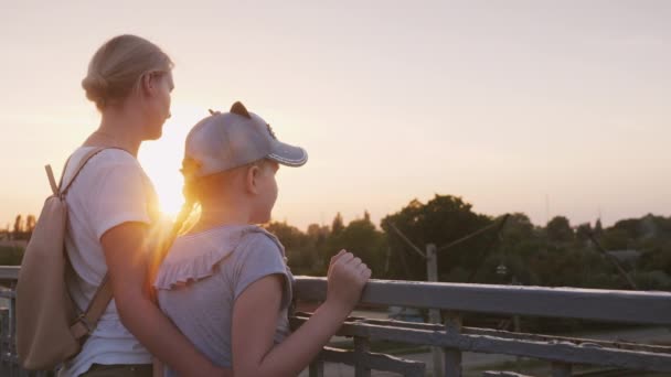 Una mujer con su hija está de pie en la barandilla del puente al atardecer. Paseo nocturno con mamá — Vídeos de Stock