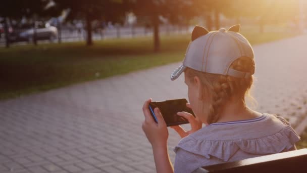 Uma menina joga no smartphone, senta-se no parque em um banco — Vídeo de Stock