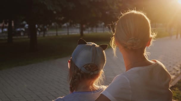 Жінка з дитиною сидить поруч на лавці в парку, перехожі йдуть — стокове відео