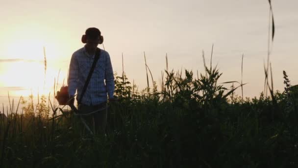 Um jovem corta a grama à noite ao pôr do sol perto de sua casa — Vídeo de Stock