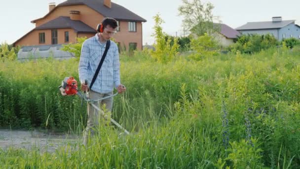 男はトリマーで家の周りに雑草を刈る — ストック動画