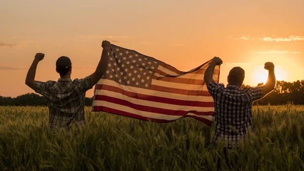 Zwei Männer hissen bei Sonnenuntergang fröhlich die amerikanische Flagge über einem Weizenfeld. 4. Juli Konzept — Stockfoto