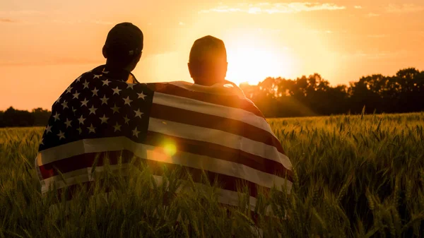 Ein Mann und sein Sohn bewundern den Sonnenuntergang über einem Weizenfeld, eingehüllt in die Flagge der USA — Stockfoto