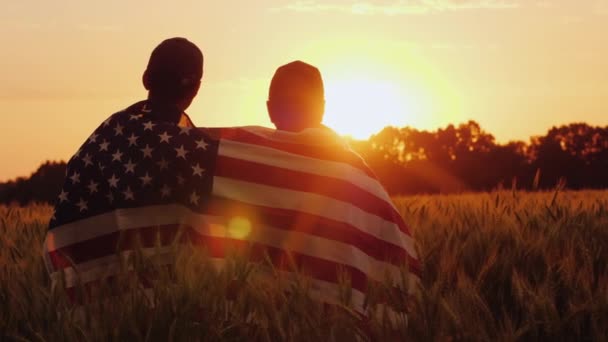 Un hombre y su hijo admiran la puesta de sol sobre un campo de trigo, envuelto en la bandera de los EE.UU. — Vídeo de stock