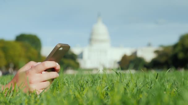 Vrouwelijke handen met een smartphone. Op het gazon, tegen de achtergrond van het Capitool in Washington. Reizen en studeren in het USA concept — Stockvideo