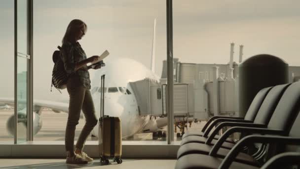 Egy nő sziluettje, akinek a beszállókártyája a terminál ablakában áll. Az ablak előtt egy gyönyörű repülőgép. — Stock videók