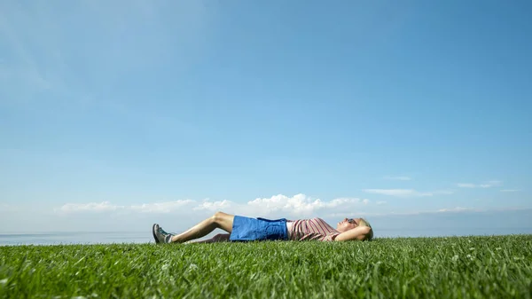 A young woman enjoys warmth and summer, lying on the green grass against the blue sky — Stock Photo, Image