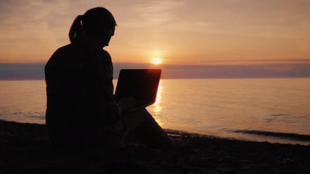 Silueta de una mujer que trabaja con un portátil junto al mar al atardecer — Vídeo de stock