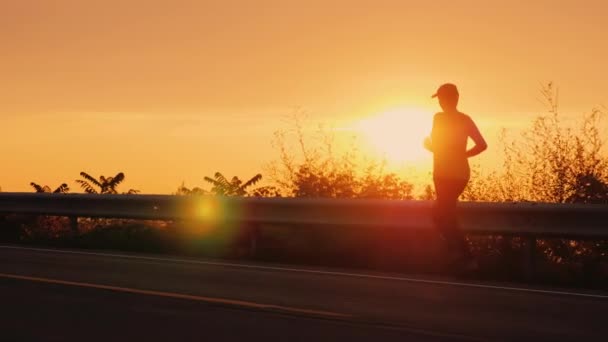 Při západu slunce vede po silnici podél pobřeží mladá žena. Silueta nerozpoznatelného člověka — Stock video