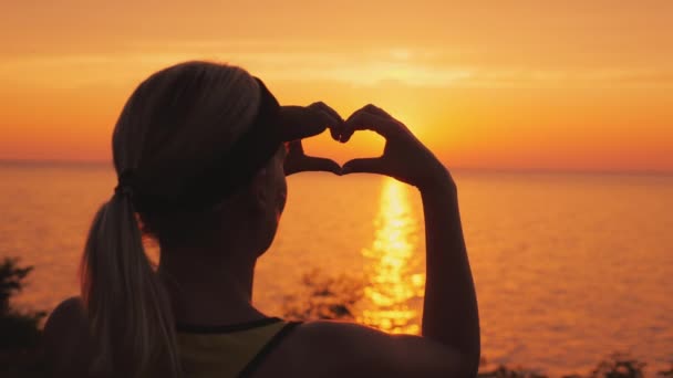 A woman looks at the sea where the sun sets, shows a heart-shaped figure — Stock Video