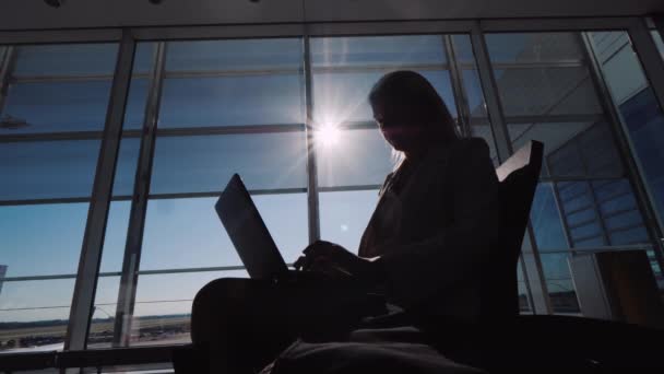 Solen belyser silhuetten av en kvinna i flygplatsterminalen, arbetar med en bärbar dator i väntan på sitt flyg — Stockvideo