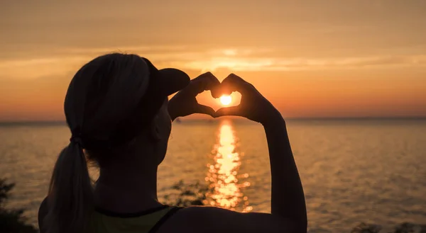 Eine Frau blickt auf das Meer, wo die Sonne untergeht, zeigt eine herzförmige Figur — Stockfoto