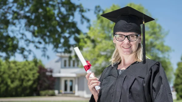 Bir manto ve mezuniyet şapkalı bir kadının portresi, evinin arka planına karşı elinde bir diploma tutan — Stok fotoğraf