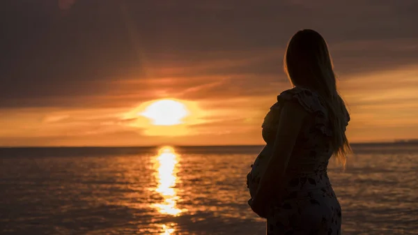 Mladá těhotná žena s velkým břichem hledí na majestátní západ slunce nad mořem. V očekávání dítěte — Stock fotografie