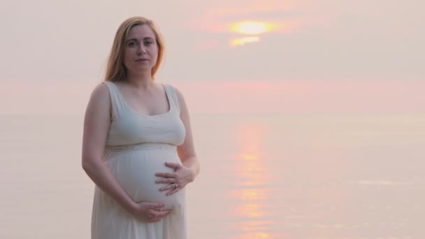 Een jonge zwangere vrouw staat in de buurt van de zee waar de zon ondergaat — Stockvideo