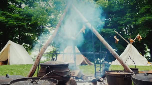 Лагерь коренных американцев в лесу. Еда готовится на переднем плане, сзади видны традиционные палатки из вигвама — стоковое видео