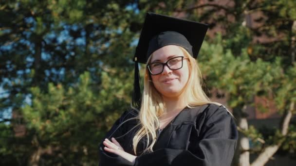 Портрет молодого випускника коледжу в одязі та шапці для випускників. Посміхаючись, дивлячись на камеру — стокове відео