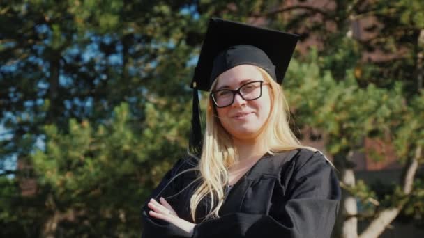 Retrato de un joven graduado universitario en ropa y una gorra de graduación. Sonriendo, mirando a la cámara — Vídeos de Stock