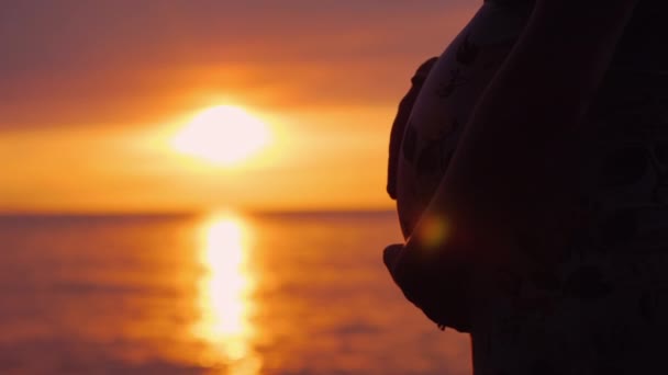 Vientre de una mujer embarazada sobre un fondo de una hermosa puesta de sol en el mar. Maternidad y esperar un concepto de bebé — Vídeo de stock