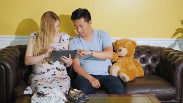 Jonge multi-etnische paar bestellingen kleren voor een ongeboren baby, gebruik een Tablet — Stockvideo