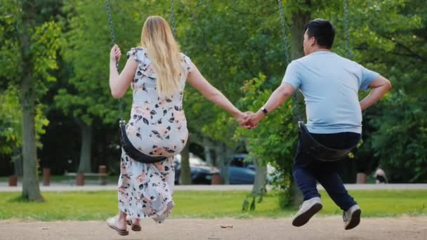 Вид сзади на мужа с беременной женой катается на качелях, держась за руки — стоковое видео