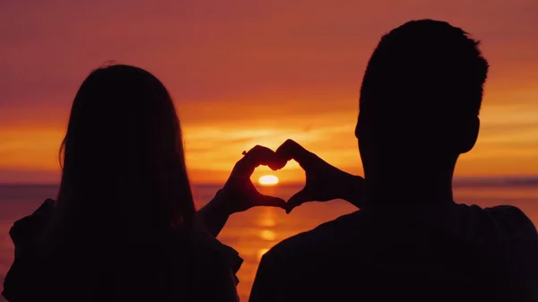 Siluety mladého páru, ruce ukazují tvar srdce na pozadí západu slunce nad mořem — Stock fotografie