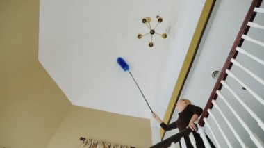 Bir kadın avizenin yanındaki tavandaki örümcek ağlarını ve enkazı temizliyor. Büyük ev temizliği